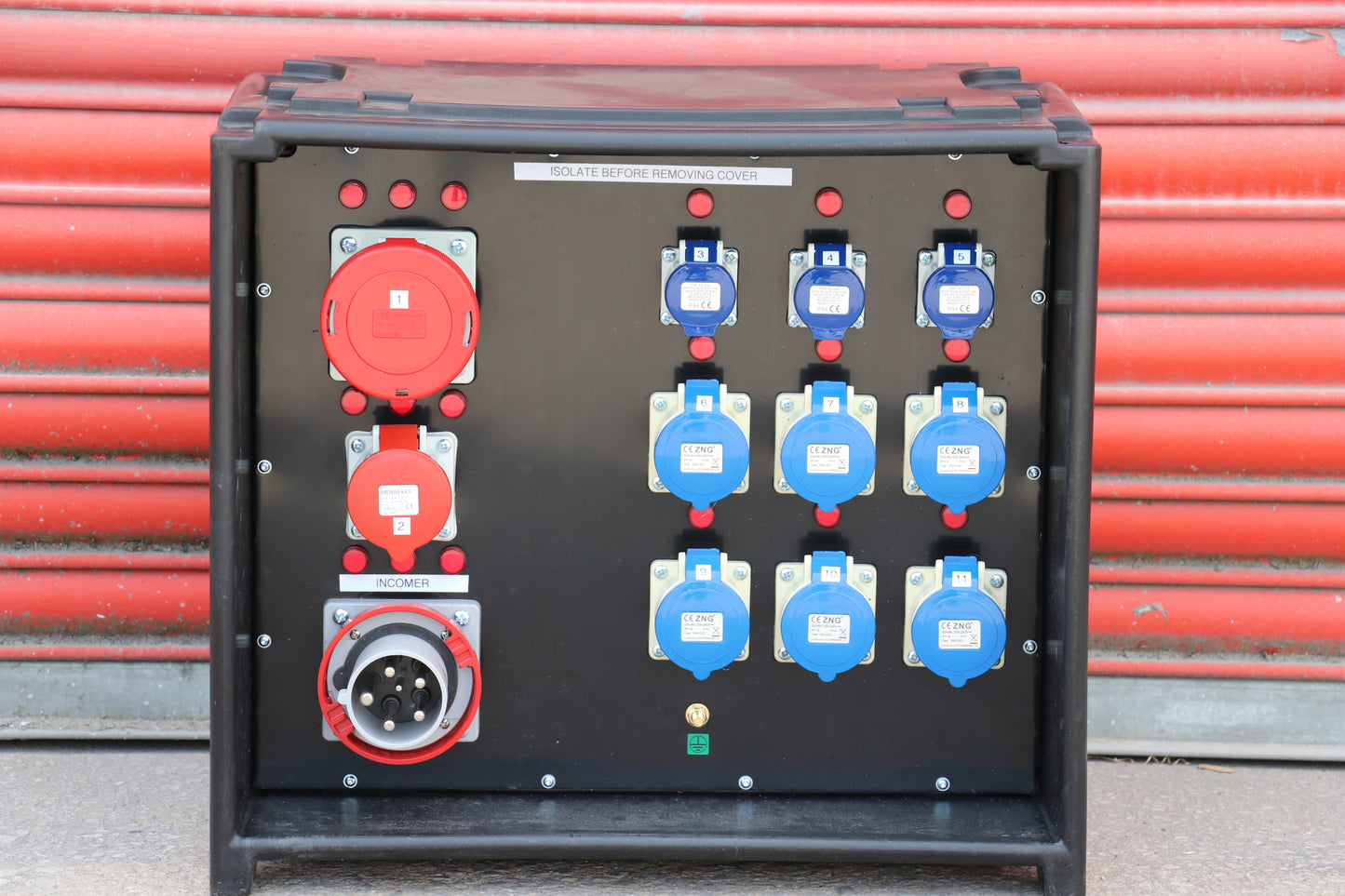 A heavy-duty black electrical distribution panel or control cabinet with multiple red and blue industrial power outlets, set against a red corrugated metal background.