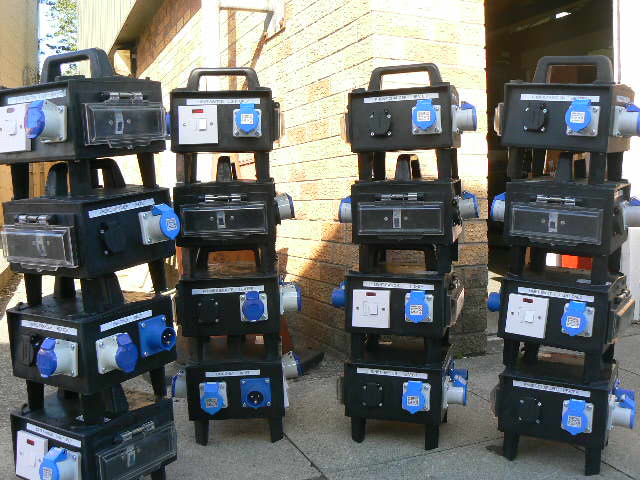 Four stacks of heavy-duty black electrical distribution cabinets with blue and white outlets, photographed outdoors against a brick wall.