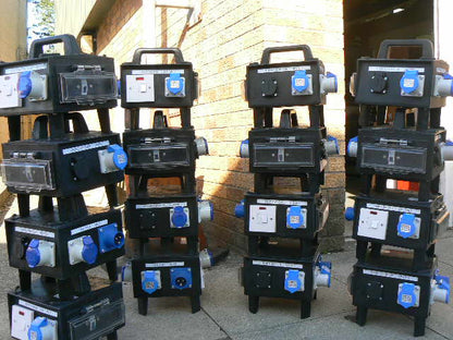 Four stacks of heavy-duty black electrical distribution cabinets with blue and white outlets, photographed outdoors against a brick wall.